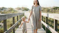 Smiling Caucasian mother in linen dress walks with toddler on sunny beach boardwalk, holding hands, sand dunes in background.