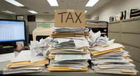 Overwhelmed office worker buried under massive pile of tax documents and paperwork in corporate accounting department during busy season.