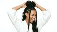 Young woman with dark skin tone and black braided hairstyle wearing white casual sweater, smiling while adjusting her hair with both hands in a joyful pose for lifestyle and beauty content.