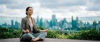 Professional businesswoman meditating in lotus pose on urban rooftop terrace with city skyline background, practicing mindfulness and wellness during work break.