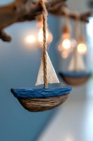 Handcrafted wooden sailboat ornament with white fabric sail hanging on twine against blurred bokeh lights, creating dreamy nautical home decor atmosphere.