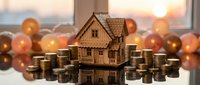 Wooden house model surrounded by coin stacks on reflective surface with warm bokeh lights, representing real estate investment and property financing concepts.