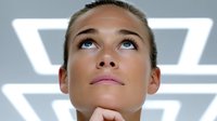 Young Caucasian businesswoman looking up thoughtfully with hand on chin in modern office with geometric lighting for corporate planning concepts.