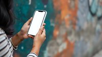 Person with medium skin tone holding smartphone with blank white screen against colorful urban graffiti wall background, suitable for mobile app mockups and technology marketing.