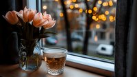 Fresh peach tulips in glass vase beside beverage on windowsill with evening city lights bokeh background, suitable for home decor and lifestyle content.