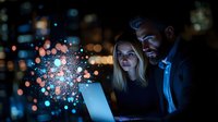 Young diverse business couple working late on laptop with magical glowing light effect and bokeh city lights in background creating atmospheric night scene.