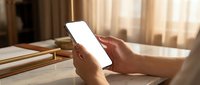 Person using smartphone with blank white screen in cozy bedroom setting with warm natural lighting and books on bedside table for mockup design.