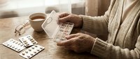 Senior woman taking medication pills from blister pack at wooden table with coffee cup, healthcare routine for elderly patient managing daily treatment.