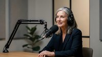 Professional mature woman with gray hair wearing headphones speaking into microphone at podcast recording studio for media content creation.