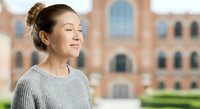 Young East Asian woman wearing gray knit sweater standing outdoors near historic brick building, smiling while looking away, suitable for lifestyle and portrait use.