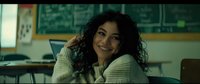 Young Hispanic woman with curly dark hair smiling warmly while sitting at desk in classroom, wearing cozy cream sweater, natural lighting creates welcoming educational atmosphere.