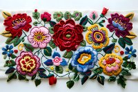 Vibrant floral embroidery pattern featuring colorful roses, sunflowers and folk art style blooms on white fabric with intricate stitching and detailed leaf elements.