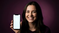 Young brunette woman smiling while holding smartphone with blank white screen against purple background for mobile app mockup and technology presentation.