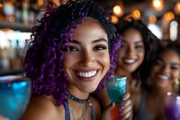 Young African American woman with vibrant purple curly hair smiling brightly at nightclub party, holding cocktail with friends in background. Joyful celebration atmosphere.