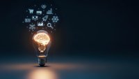 Glowing light bulb with brain inside and business icons floating above on dark blue background representing innovation and creative thinking.
