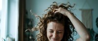 Young woman with curly hair laughing while running hand through hair in bright indoor setting. Natural joy and carefree lifestyle moment.