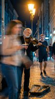 Senior street musician playing violin on cobblestone evening street with warm lamplight and blurred pedestrians walking by in urban setting.
