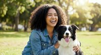 Happy young African American woman laughing while hugging her Border Collie dog in sunny park. Joyful pet owner bonding with furry companion outdoors.