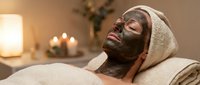 Young woman relaxing with dark clay facial mask and towel on head in cozy spa salon with candles, enjoying skincare treatment.