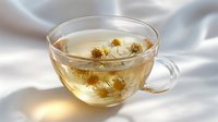Glass cup of hot chamomile tea with dried flowers floating on surface against white silk fabric background, soft natural lighting creates gentle shadows.