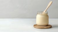 Creamy homemade mayonnaise in glass jar with wooden spoon on round bamboo coaster against light background, minimalist food photography styling.