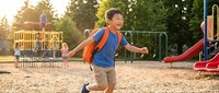 Happy Asian elementary school boy with backpack running excitedly at playground during golden hour sunset with colorful equipment and trees in background.