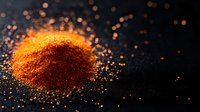 Vibrant red chili powder heap with scattered spicy particles on dark background, dramatic food photography with orange bokeh effect for culinary designs.