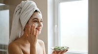 Young woman applying natural skincare cream in bright bathroom with towel wrapped hair for daily beauty routine and self-care wellness lifestyle.