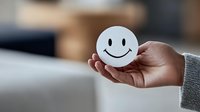 Hand in gray sweater sleeve holding white round card with classic smiley face drawing, representing happiness, positivity and emotional wellbeing concept.