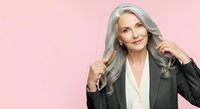 Mature woman with long gray hair wearing black blazer and white blouse, posing against pink background. Suitable for business, lifestyle, and age-positive marketing campaigns.