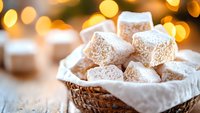 Powdered sugar coated Turkish delight cubes in woven basket with festive bokeh lights background, soft focus close up view creates cozy holiday atmosphere.