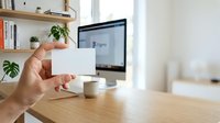 Male hand holding blank white business card at wooden desk with computer monitor, potted plant, and shelving in bright office workspace for corporate branding and professional identity presentation.