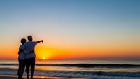 Romantic couple silhouette embracing on beach at golden sunset, man pointing toward horizon over ocean waves, peaceful evening moment together.