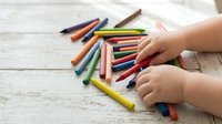 Child hands selecting colorful crayons from scattered collection on white wooden table for creative art project and drawing activities.