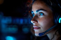 Young female gamer with freckles wearing gaming headset in dark room illuminated by blue light, side profile view showing focused expression and glowing screen reflection.