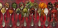 Colorful array of aromatic spices and herbs in vintage silver spoons arranged on dark red background, overhead view showing diverse seasonings and flavors.