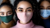 Young multiracial women wearing protective face masks looking at camera, focus on middle eastern woman in pink mask and sweater during pandemic prevention.