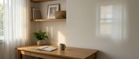Minimalist home office workspace with wooden desk, floating shelf, plant and natural sunlight streaming through window creating warm shadows on wall.