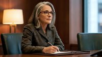 Mature woman with gray hair and glasses wearing gray blazer sitting at desk in modern office, professional business setting.
