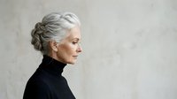 Mature woman with silver gray hair styled in elegant updo, wearing black turtleneck, profile view against neutral background. Suitable for aging, beauty, fashion, and lifestyle editorial use.