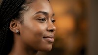 Young African American woman with braided hair and earrings smiling confidently in warm indoor lighting for lifestyle and portrait photography.