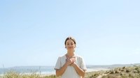 Woman with blonde hair, Caucasian ethnicity, wearing glasses and casual beige shirt, standing outdoors in natural landscape with ocean and hills visible behind her, hands clasped together in thoughtful pose.
