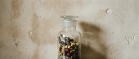 Dried herbs and flowers stored in glass jar with cork lid against textured beige wall, suitable for herbal medicine, wellness, and natural remedy packaging design.