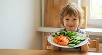 Happy blonde toddler boy sitting at wooden table holding white plate with fresh vegetables including carrots and cucumbers in bright kitchen interior.