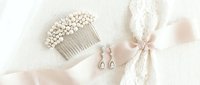 Elegant bridal accessories set with pearl hair comb, drop earrings and lace ribbon on soft pink silk fabric for wedding day preparation.