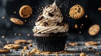 Chocolate cupcake with cream cheese frosting swirled into peaks, topped with cookie crumbles and chocolate chips against dark background with flying crumbs.