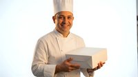 Middle-aged male chef of South Asian ethnicity wearing white chef's coat and tall chef's hat, smiling while holding an open white box against white background.