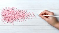 Female hand with pencil drawing flying pink paper hearts on white wooden background, romantic creative concept for Valentine's day cards and love messages.