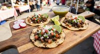 Three authentic Mexican tacos with meat, onions, cilantro and lime on wooden board at busy street food market. Traditional Latin American cuisine ready to serve.