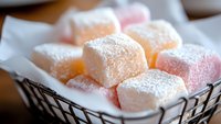 Pink and peach Turkish delight cubes dusted with powdered sugar nestled in white paper inside dark wire basket, macro food photography with soft focus background.